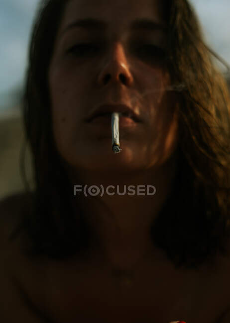 D'en bas flou brun bouclé femme aux cheveux regardant caméra avec cigarette dans la bouche — Photo de stock