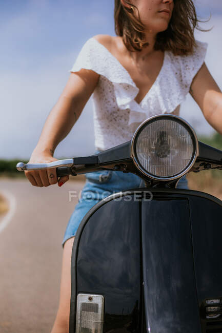 Обрезать молодую женщину в повседневной одежде вождения скутера на дороге в солнечный день — стоковое фото