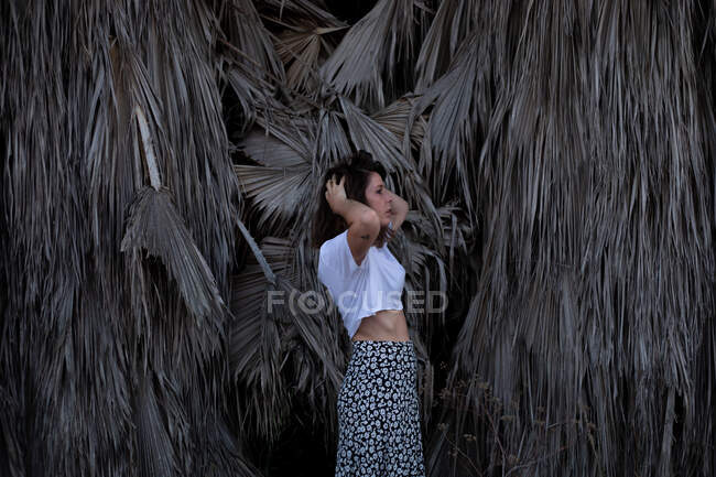 Side view of young brunette in white t shirt and light skirt standing and fixing hair with tropical dry plants on background — Stock Photo