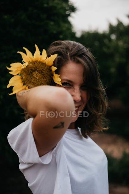 Side view of brown haired woman in light clothing looking at camera holding sunflower behind ear amid green trees during summer vacation — Stock Photo