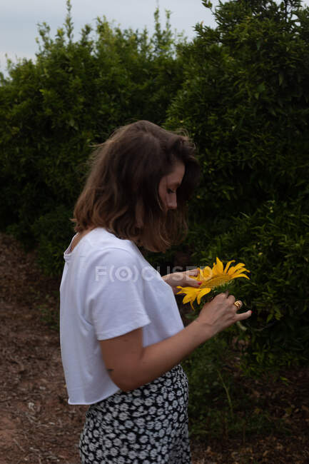 Vue latérale de la femme aux cheveux bruns en vêtements légers avec debout avec fleur dans les mains au milieu des arbres verts pendant les vacances d'été — Photo de stock