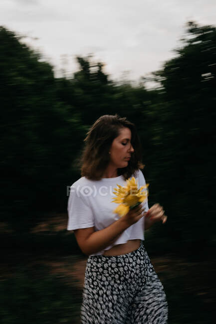 Vista lateral de la mujer de cabello castaño en ropa ligera con pie con flor en las manos en medio de árboles verdes durante las vacaciones de verano mirando a la cámara - foto de stock