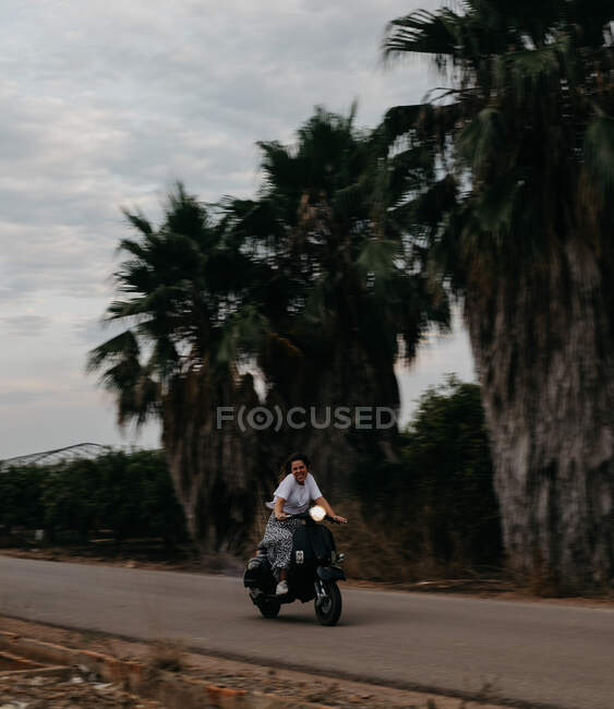 Satisfied woman on vacation in light clothing smiling and looking at camera on motorcycle in motion with tropical plants on background — Stock Photo