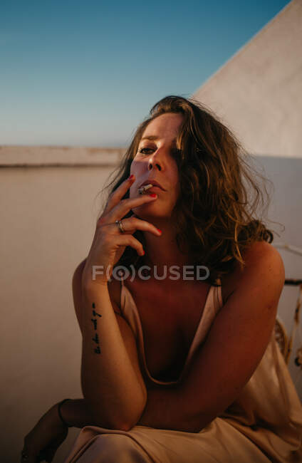 Mujer pacífica sentada mirando hacia otro lado cerca de la cerca del balcón y fumando cigarrillo con puesta de sol sobre fondo borroso - foto de stock