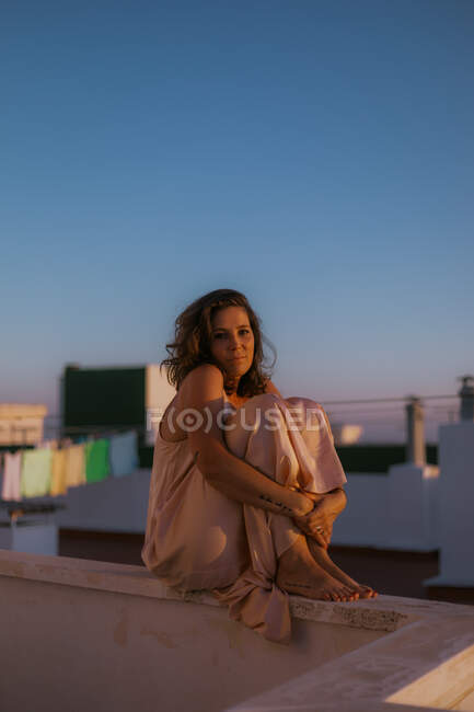 Joven mujer de pelo rizado en elegante vestido de seda rosa sentado en la cerca del balcón con los brazos alrededor de las piernas y mirando a la cámara durante el atardecer de verano - foto de stock