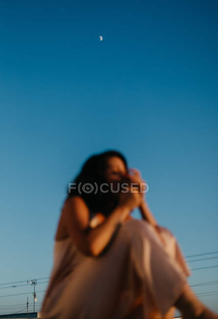 Blurred young woman in stylish pink silk dress sitting on fence of balcony smoking cigarette during summer sunset — Stock Photo