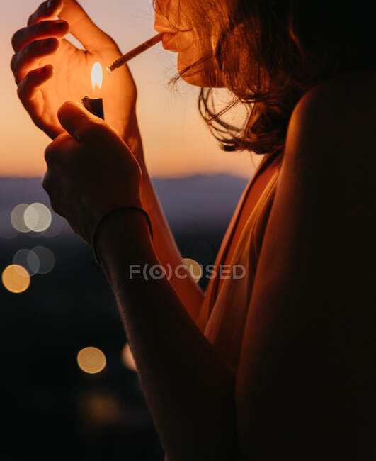 Vue latérale de la femme cultivée allumant cigarette avec magnifique coucher de soleil sur fond flou — Photo de stock