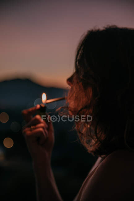 Vista lateral da jovem mulher sem rosto acendendo cigarro com magnífico pôr do sol no fundo embaçado — Fotografia de Stock