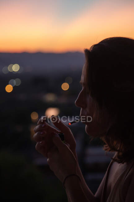 Vista lateral da jovem mulher acendendo cigarro com magnífico pôr do sol no fundo embaçado — Fotografia de Stock