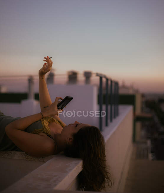 Вид збоку жінки, що лежить на паркані балкона, курить сигарету і використовує мобільний телефон, щоб сфотографуватися із заходом сонця на розмитому тлі — стокове фото