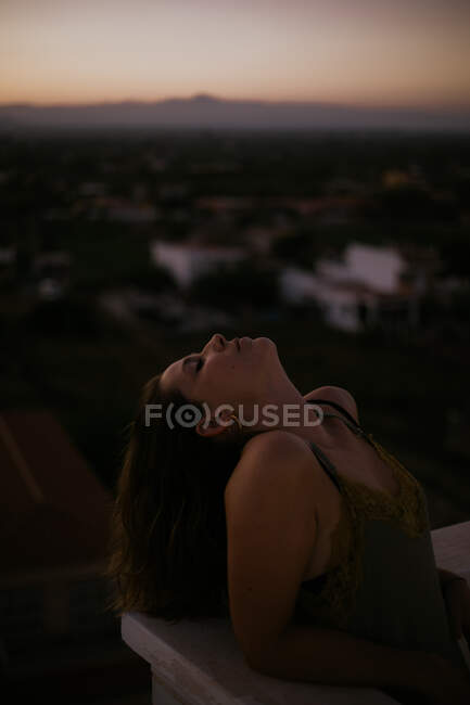 Mulher pacífica inclinada com os olhos fechados na cerca da varanda com o pôr do sol no fundo embaçado — Fotografia de Stock