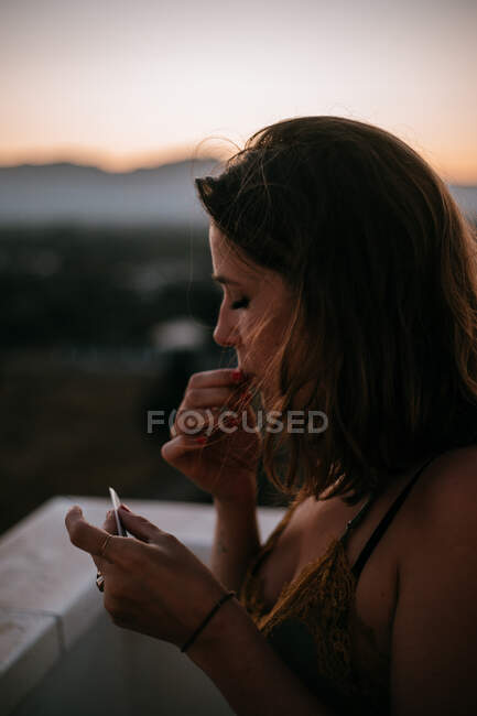 Vista lateral de la mujer de pie con los ojos cerrados cerca de la cerca del balcón y la preparación de cigarrillo para fumar con puesta de sol sobre fondo borroso - foto de stock