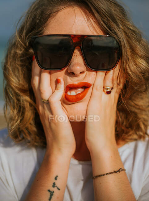 Стильная каштановая кудрявая женщина с красной помадой в модных солнцезащитных очках, смотрящая в камеру, сжимая лицо в ладонях — стоковое фото