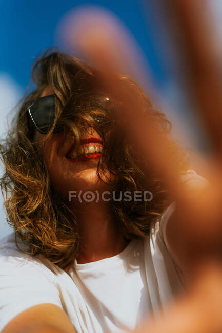 Von unten stylische Frau mit braunen Locken und rotem Lippenstift in trendiger Sonnenbrille, die in die Kamera mit blauem Himmel auf dem Hintergrund blickt — Stockfoto