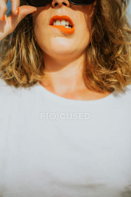 Elegante mujer de pelo rizado marrón con lápiz labial rojo en gafas de sol de moda mirando a la cámara mientras hace caras - foto de stock