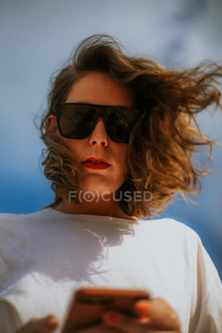 Снизу стильная женщина на отдыхе в белой футболке модные солнцезащитные очки и красные губы глядя и камеры при использовании мобильного телефона с голубым небом на заднем плане — стоковое фото
