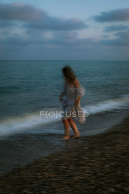 Barefoot female traveler in light dress dancing among small sea waves on empty coastline at dusk — Stock Photo