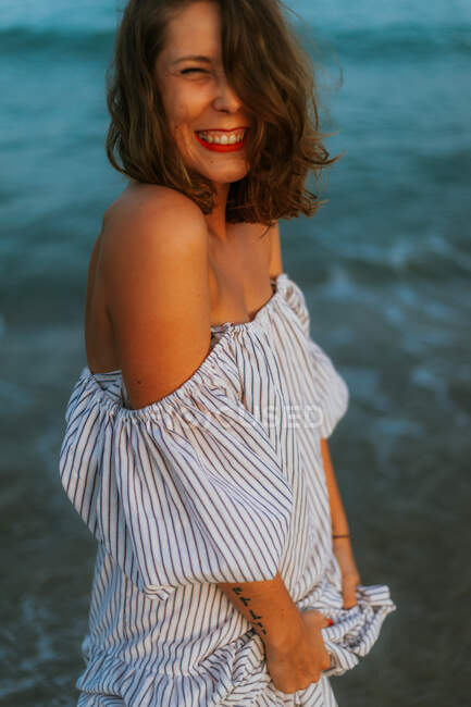 Mulher feliz em vestido de luz andando entre pequenas ondas do mar na costa vazia ao entardecer com os olhos fechados — Fotografia de Stock