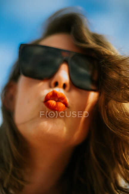 De dessous élégant brun bouclé femme aux cheveux avec rouge à lèvres dans des lunettes de soleil à la mode en regardant la caméra avec ciel bleu sur le fond — Photo de stock