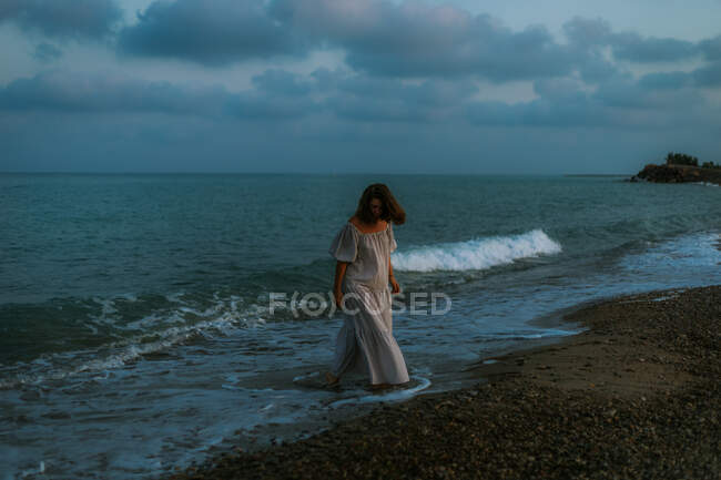 Viaggiatrice a piedi nudi in abito leggero che cammina tra piccole onde marine sulla costa vuota al crepuscolo guardando altrove — Foto stock