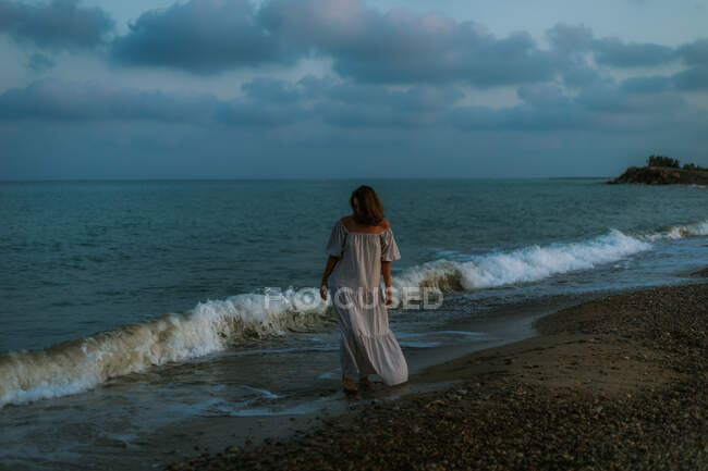 Descalço viajante feminino em vestido leve andando entre pequenas ondas do mar na costa vazia ao anoitecer olhando para longe — Fotografia de Stock