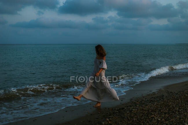 Босоногая женщина-путешественница в легком платье ходит среди маленьких морских волн на пустой береговой линии в сумерках и смотрит в сторону — стоковое фото
