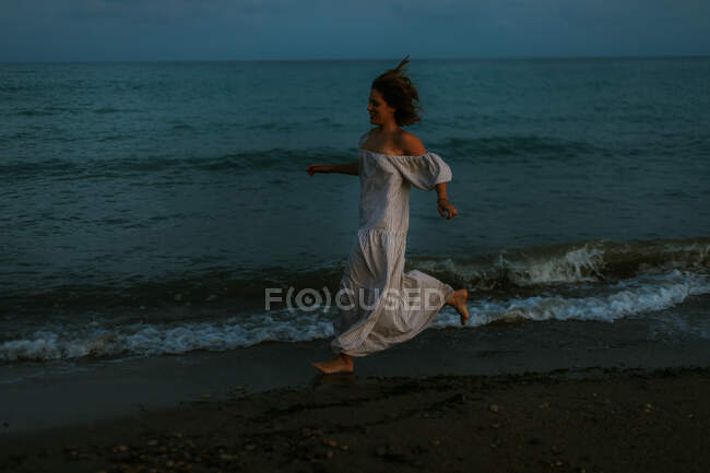 Barefoot female traveler in light dress running among small sea waves on empty coastline at dusk looking away — Stockfoto