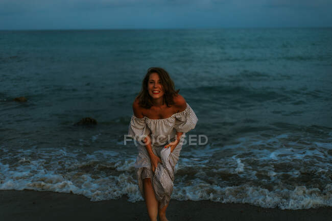Barefoot female traveler in light dress dancing among small sea waves on empty coastline at dusk looking at camera — Stock Photo