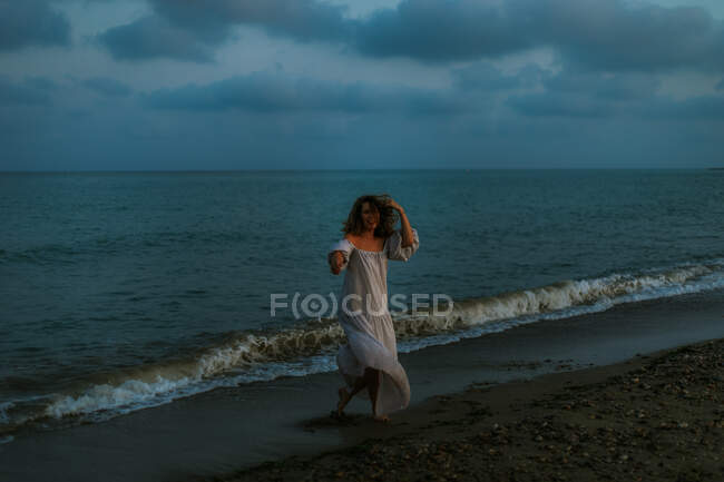 Босоногая женщина-путешественница в легком платье танцует среди маленьких морских волн на пустой береговой линии в сумерках, глядя в камеру — стоковое фото