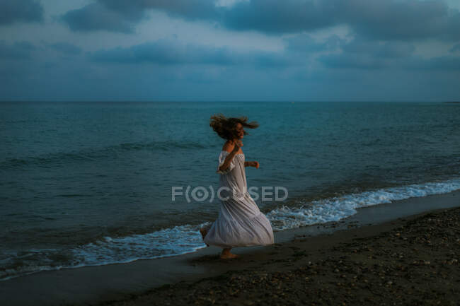 Side view anonymous of barefoot woman traveler in light dress dancing among small sea waves on empty coastline at dusk looking away — Stock Photo