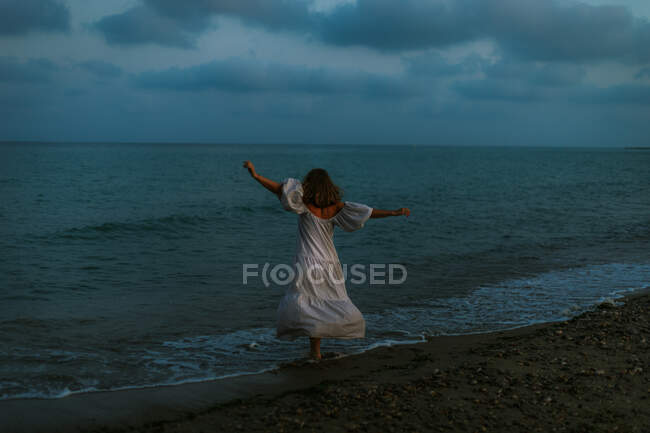 Вид ззаду анонімний босоніж жінка мандрівник у легкій сукні танцює серед невеликих морських хвиль на порожній береговій лінії в сутінках, дивлячись далеко — стокове фото
