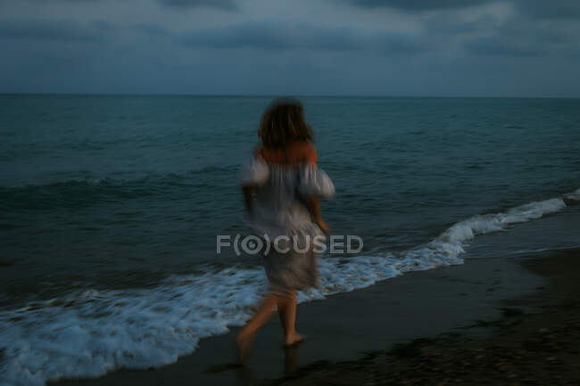 Back view of anonymous barefoot woman traveler in light dress running among small sea waves on empty coastline at dusk — Stockfoto