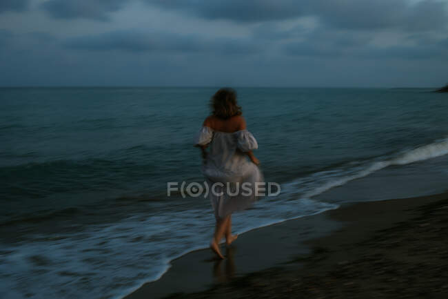 Vue de dos de voyageuse anonyme pieds nus en robe légère courant parmi les petites vagues de mer sur le littoral vide au crépuscule — Photo de stock