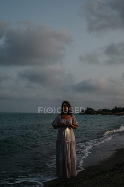 Barefoot female traveler in light dress walking among small sea waves on empty coastline at dusk looking at camera — Stockfoto