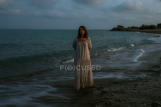 Босоногая женщина-путешественница в светлом платье ходит среди маленьких морских волн на пустой береговой линии в сумерках, глядя в камеру — стоковое фото