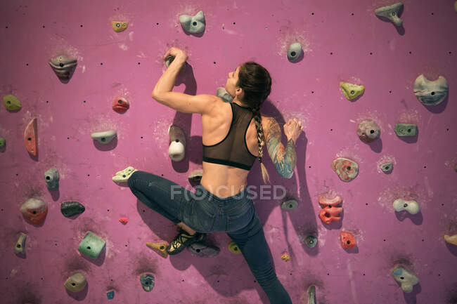 Vista posterior de atleta irreconocible tatuado poderosa mujer escalando en la pared colorida con repisas para escaladores en la habitación - foto de stock