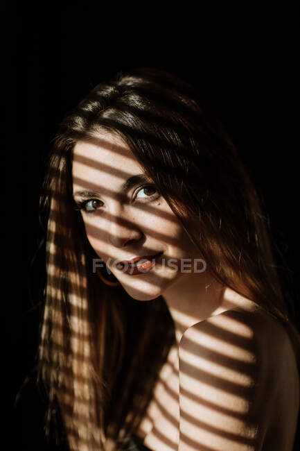Тень от жалюзи падает на лицо очаровательной расслабленной длинноволосой женщины, улыбающейся в камеру — стоковое фото