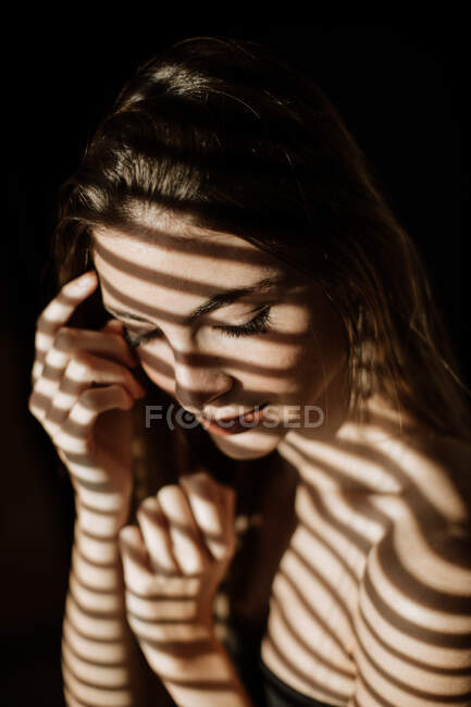 Streifenschatten von Rollläden fallen auf das Gesicht einer charmant entspannten langhaarigen Frau, die mit geschlossenen Augen lächelt — Stockfoto