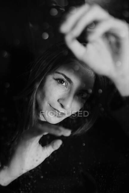 Black and white of smiling woman standing behind glass in water drops touching surface and looking at camera — Stock Photo