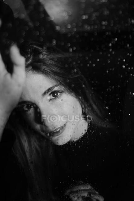 Blanco y negro de mujer sonriente de pie detrás del vidrio en gotas de agua tocando la superficie y mirando a la cámara - foto de stock