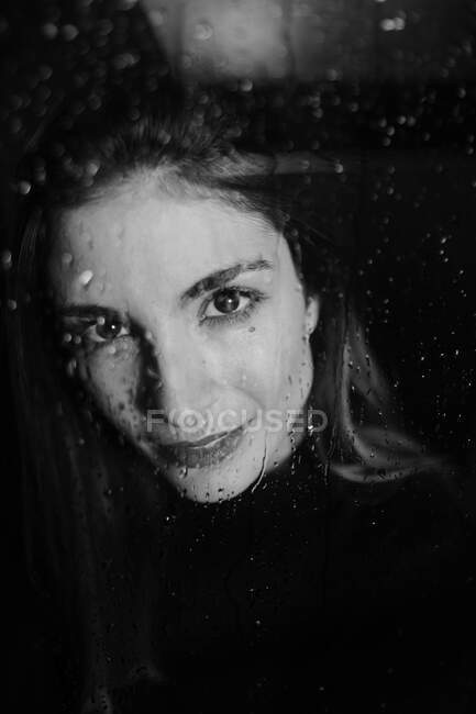 Preto e branco de mulher sorridente em pé atrás de vidro em gotas de água tocando superfície e olhando para a câmera — Fotografia de Stock