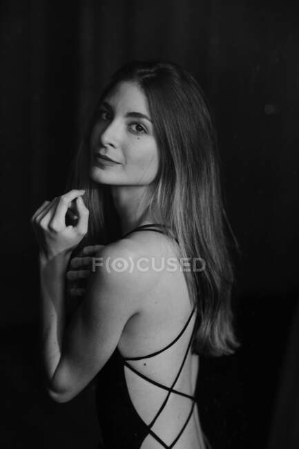 Long haired stylish woman in dark dress with with open back lacing alluringly looking over shoulder at camera on black background — Stock Photo