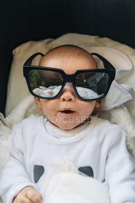 Зверху новонародженої дитини в піжамі з відкритим ротом в сонцезахисних окулярах, лежачи на ліжку вдома — стокове фото