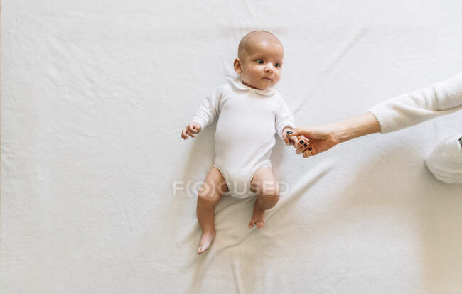 Вид сверху на безликую женщину, трогающую руки веселого новорожденного младенца с открытым ртом в белой пижаме, весело лежащего на кровати и отводящего взгляд — стоковое фото