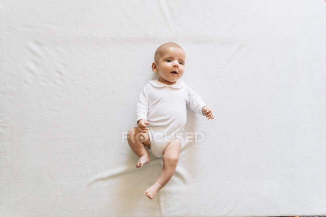 Top view of lovely infant in white pajama with open mouth lying on bed moving arms and legs looking up — Stock Photo