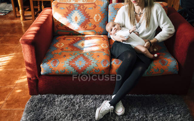Triste neonato in pigiama bianco sulle braccia della madre che si prende cura delle colture con le gambe incrociate seduto sul letto vicino ai cani a casa — Foto stock