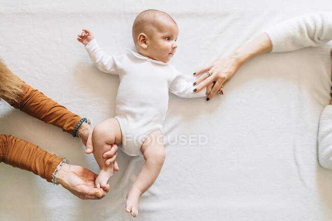 Vista superior de mujeres sin rostro tocando las manos de un alegre bebé recién nacido con la boca abierta en pijama blanco divirtiéndose tumbado en la cama mirando hacia otro lado - foto de stock