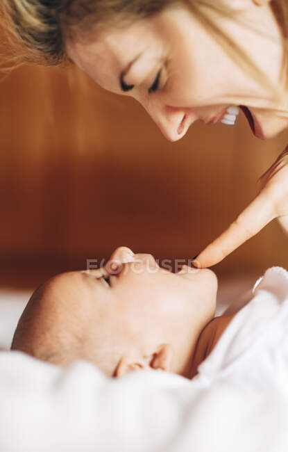 Крупный план радостной мамы, касающейся носа за носом ребенка, играющей с новорожденным младенцем с открытым ртом, веселящимся лежа на кровати дома — стоковое фото