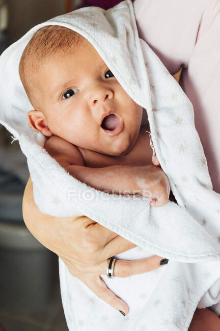 Closeup of calm newborn infant in white blanket on arms of crop caring mother at home looking at camera — Stock Photo