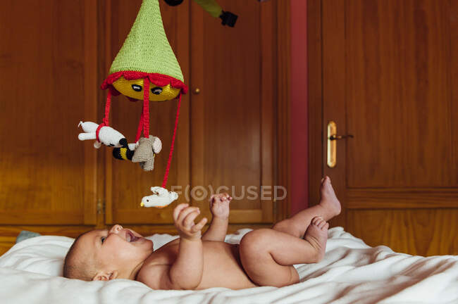 Seitenansicht des fröhlichen nackten Babys mit offenem Mund, das mit Spielzeug spielt, das auf dem Bett liegt und zu Hause Spaß hat — Stockfoto